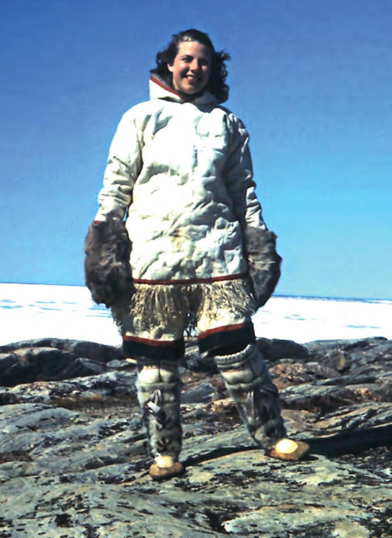 A woman stands on a rocky tundra wearing a parka and tall animal skin boots.