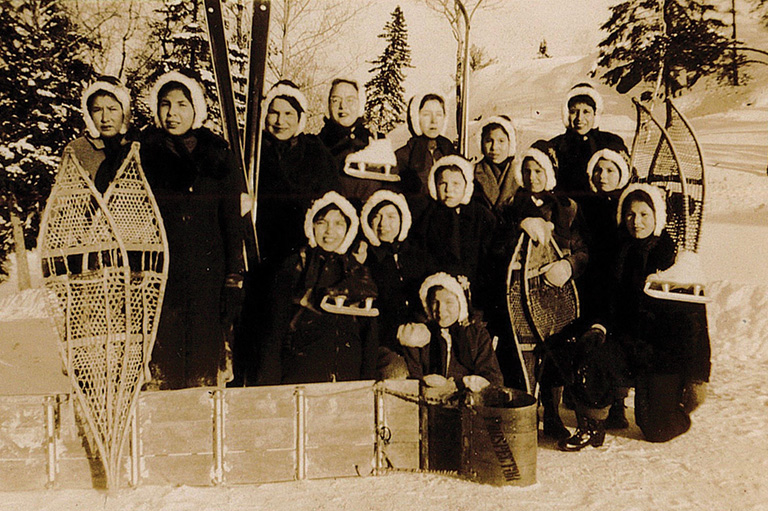 Sepia tone photo of girls in winter apparel with snow shoes and skates.