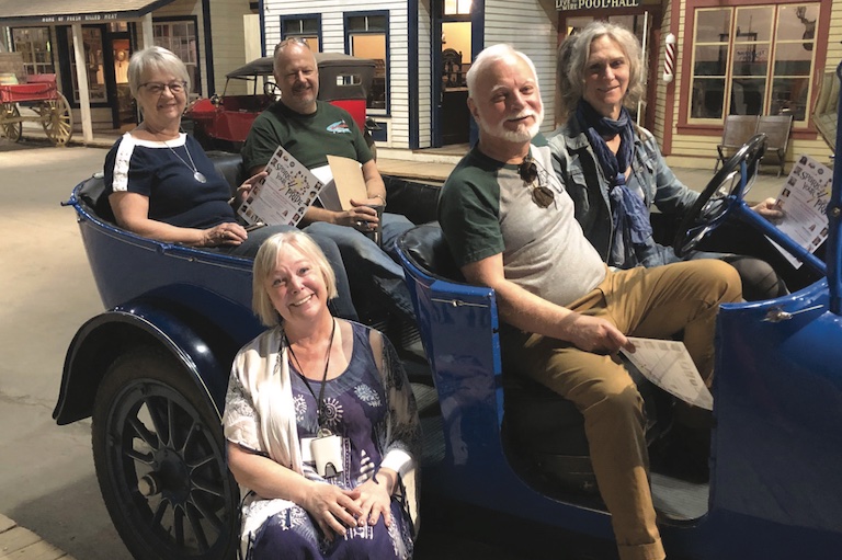 Four people sit in an old blue vehicle. One woman sits on the side step of the vehicle.