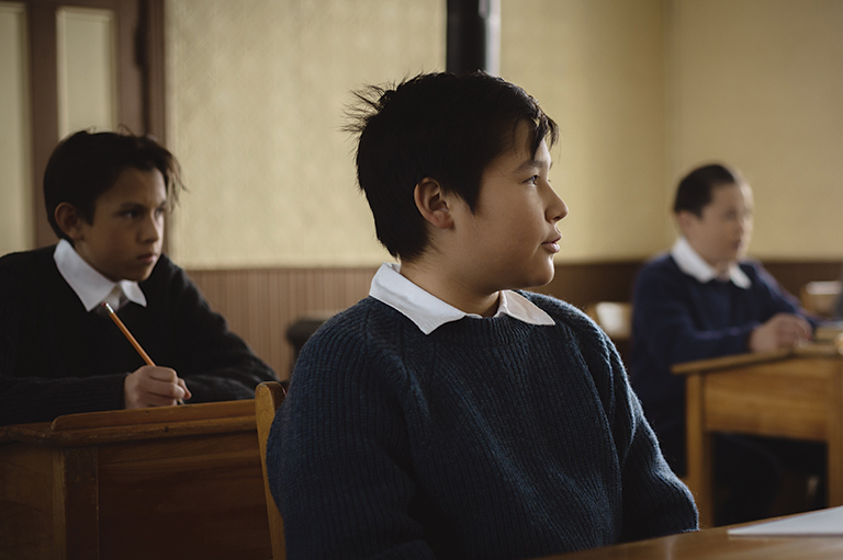 Three Indigenous boys in school uniforms, sitting at desks in a classroom.