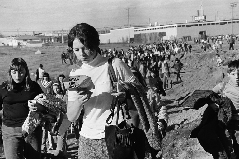 A women walks and reads while passing by the camera. Large crowd of people behind her. 