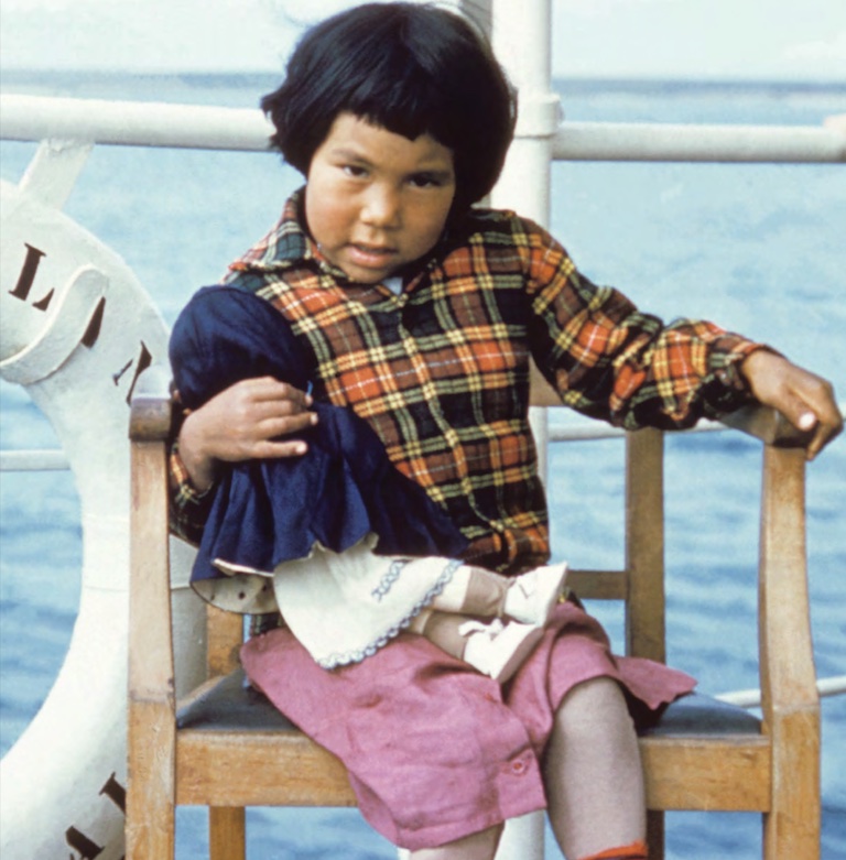 A young Inuk girl sits on a chair on a boat. She wears a plaid jacket and pink skirt and holds a doll in her hand.