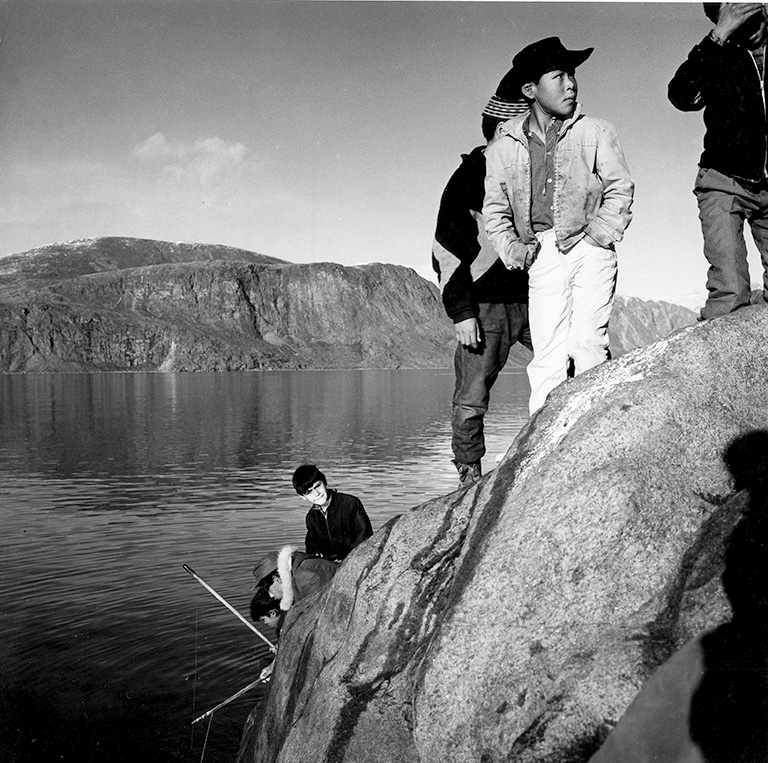 Three boys stand on the top of a rockface near the water, one wears a black cowboy hat. There are three boys sitting closer to the water who are fishing. The photo is in black and white.