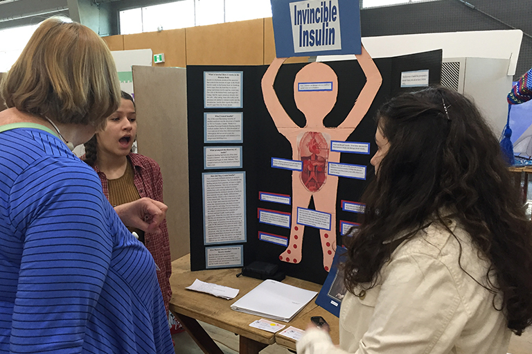 This image shows two students speaking with a judge in front of their project called "Invincible Insulin."