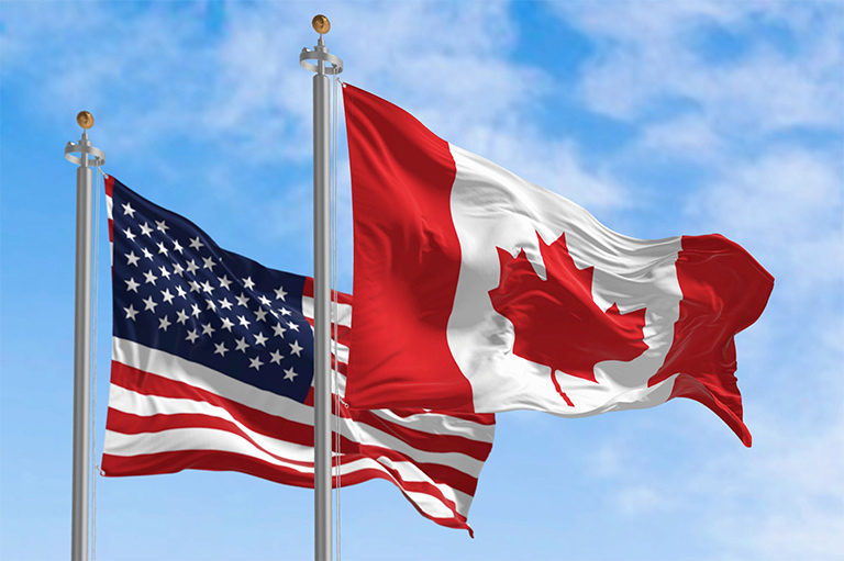 Colour photo of two waving flags: Canada in front to the right, the US on the left behind it.