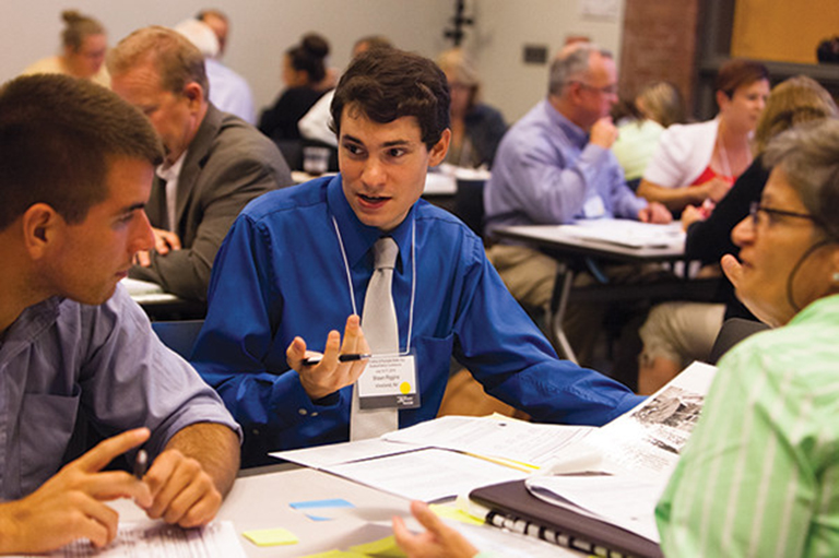 This image shows a group of educators talking around a table, on is wearing a purple shirt, one a green shirt, and one a blue shirt and a white tie. 