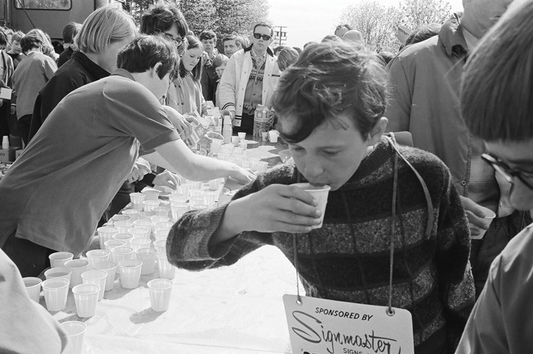 A young boy leans over the table while taking a sip of his small cup of water. 