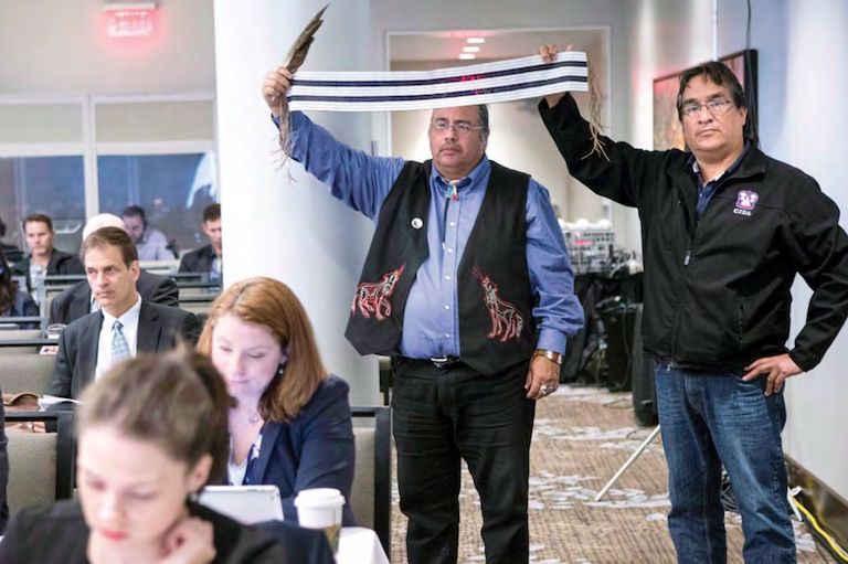 Two men stand at the side of a meeting room. They hold a long wampum belt of purple and white beads in the air between their hands. One man also grasps a feather in the hand holding the belt.