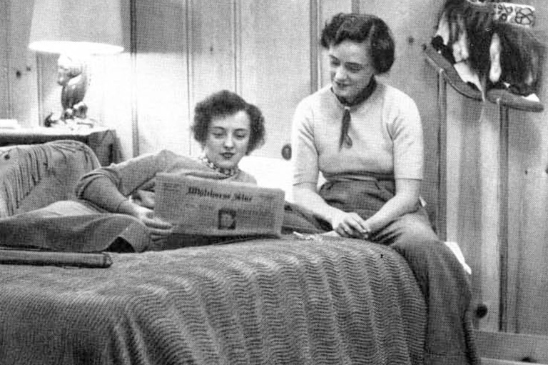 One women lays on a bed reading a newspaper, while another women sitting on the be reads over her shoulder. 