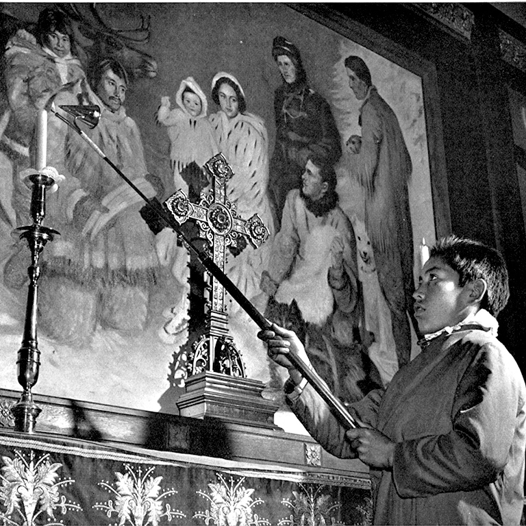 Black and white photo of a boy holding a large candle lighter in front of an altar with a mural on the wall.