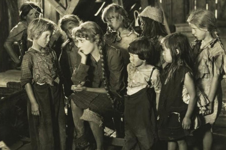 Mary Pickford sitting with her chin in her hand and elbow on her knee, surrounded by young children. 