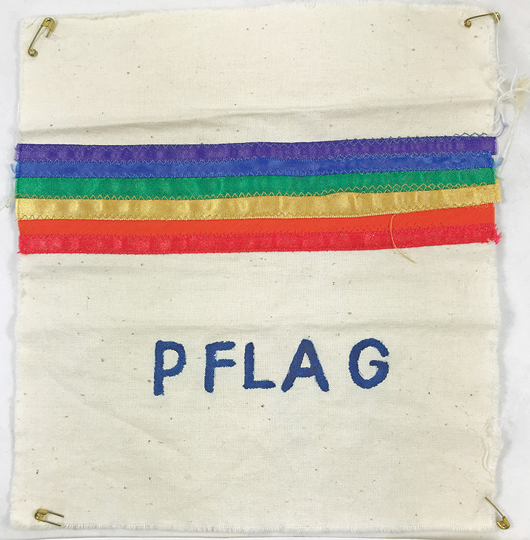 A small square cloth with safety pins in each corner has rainbow ribbons across the top and the letters PFLAG written at the centre.