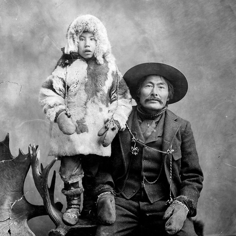 Black and white portrait of a seated man in a brimmed hat with a moustache. His young son stands beside him on a platform wearing a fur parka and mittens.