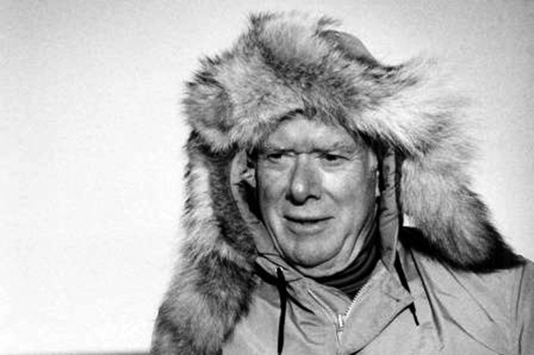 Black and white photo of a man from the shoulders up with wind blowing in his face. He wears a parka and the fur trim is being moved by the wind.