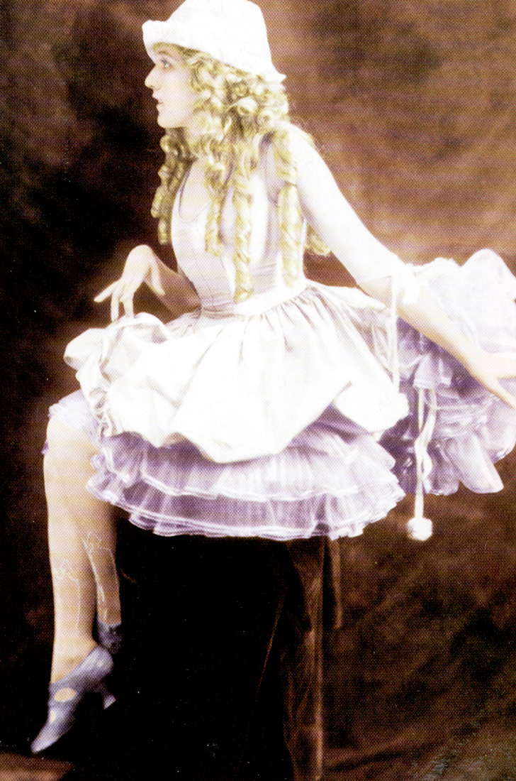Mary Pickford facing left with her whole body turned away from the camera sitting on a box, wearing a light pink dress and large curls in her hair. 