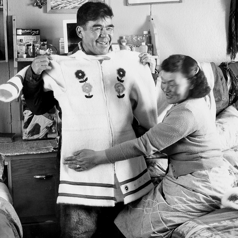 A woman stands beside a man who holds up a white parka across his chest to demonstrate the sizing.